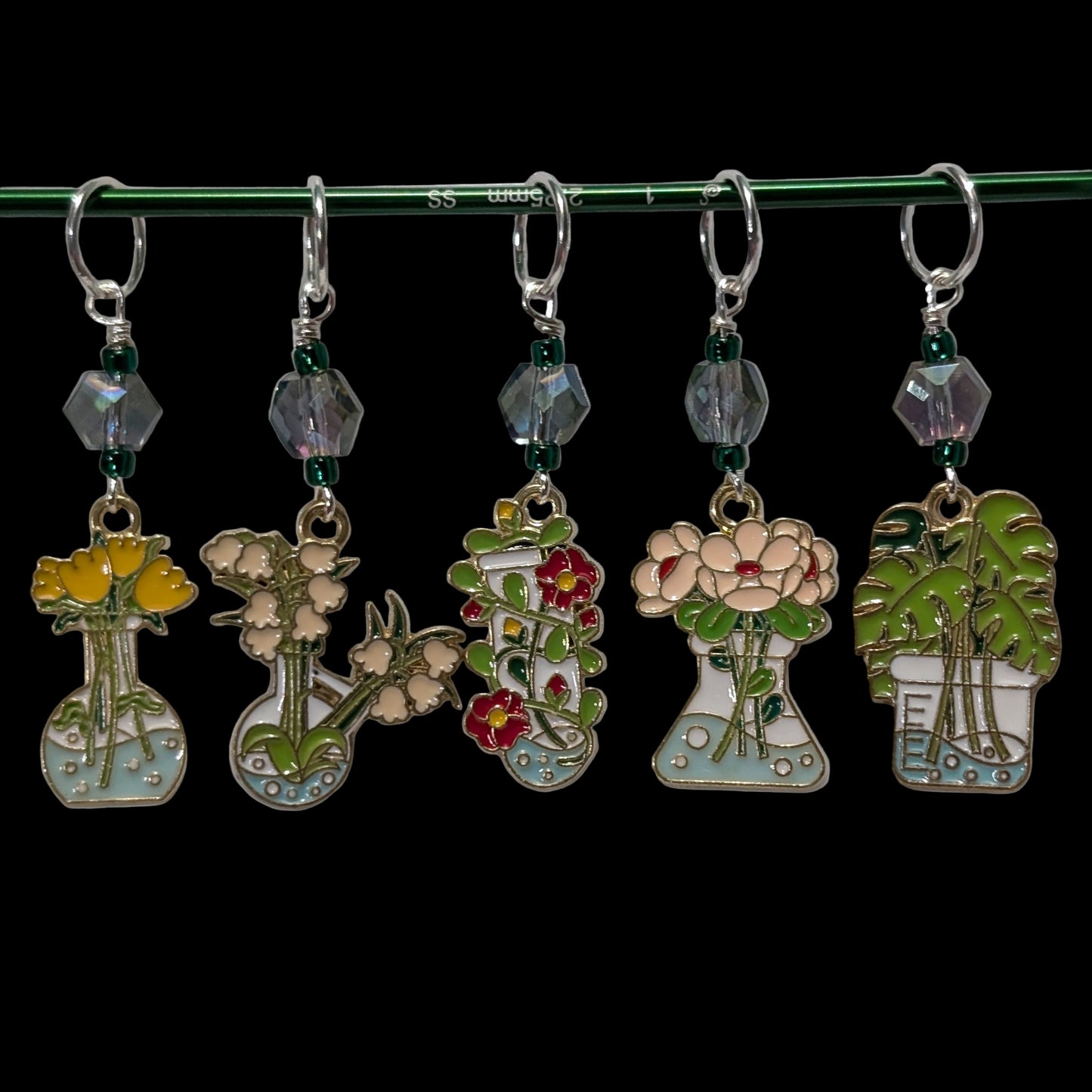 Set of five floral-themed stitch markers charms on a green rod against a black background