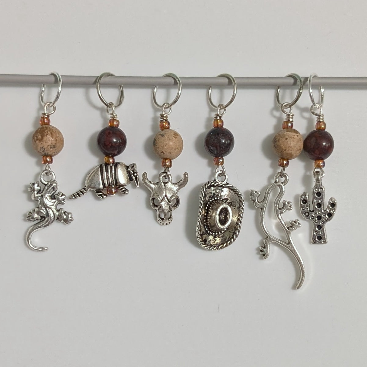 Set of six decorative stitch markers with beads and metallic elements on a white background.  Elements include 2 different lizards, an armadillo, a cow skull, a cowboy hat, and a cactus