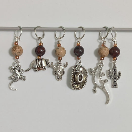 Set of six decorative stitch markers with beads and metallic elements on a white background.  Elements include 2 different lizards, an armadillo, a cow skull, a cowboy hat, and a cactus