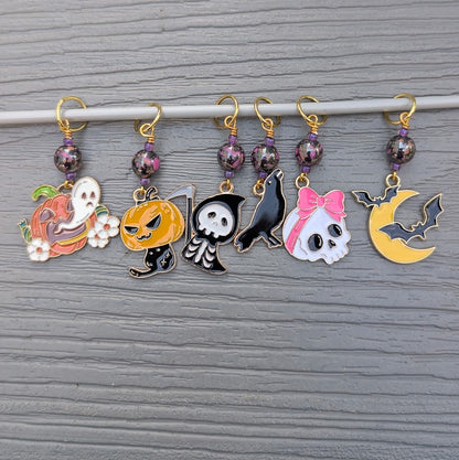 Six Halloween-themed knitting stitch markers with gold-tone rings, purple marbled beads, and colorful enamel charms hanging from a gray rod against a gray wood-textured background.