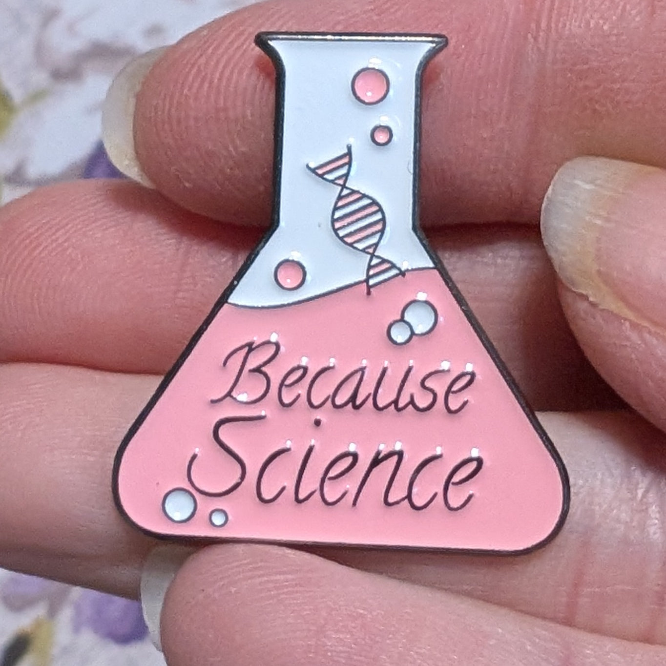 Pink beaker-shaped pin with 'Because Science' text held between fingers