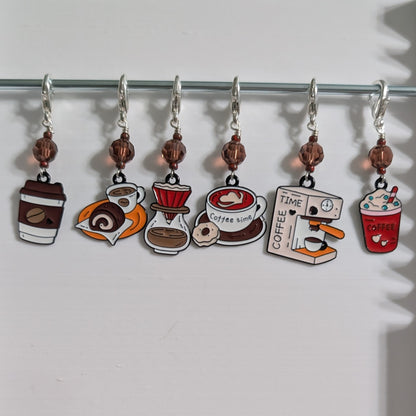 Six coffee-themed charm stitch markers hang from a silver metal rod on a white background. The charms feature illustrations of a takeaway coffee cup, a croissant, a pour-over coffee dripper, a coffee mug, a coffee machine, and a red iced coffee drink.