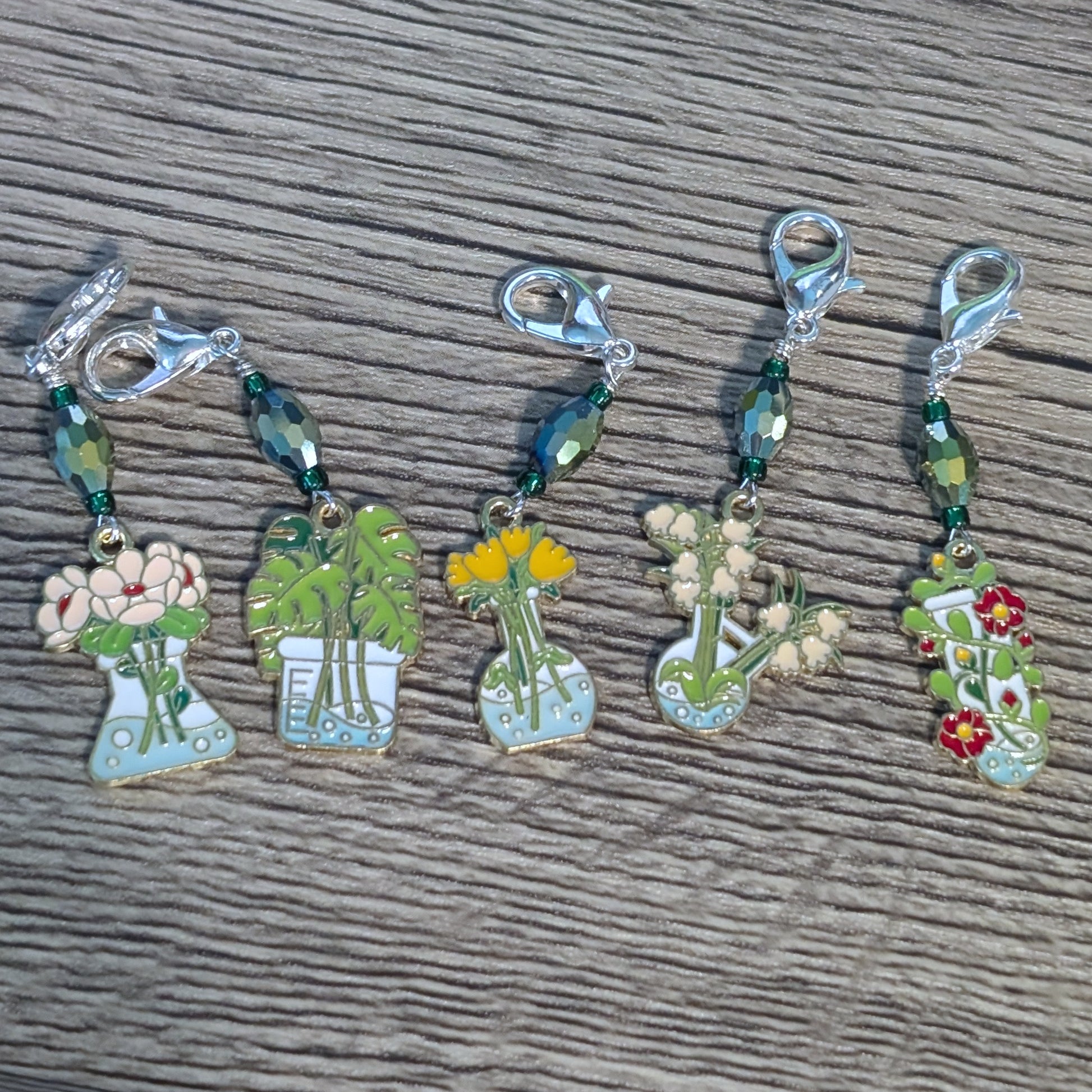 Set of five decorative stitch markers with plant and flower designs on a wooden surface.