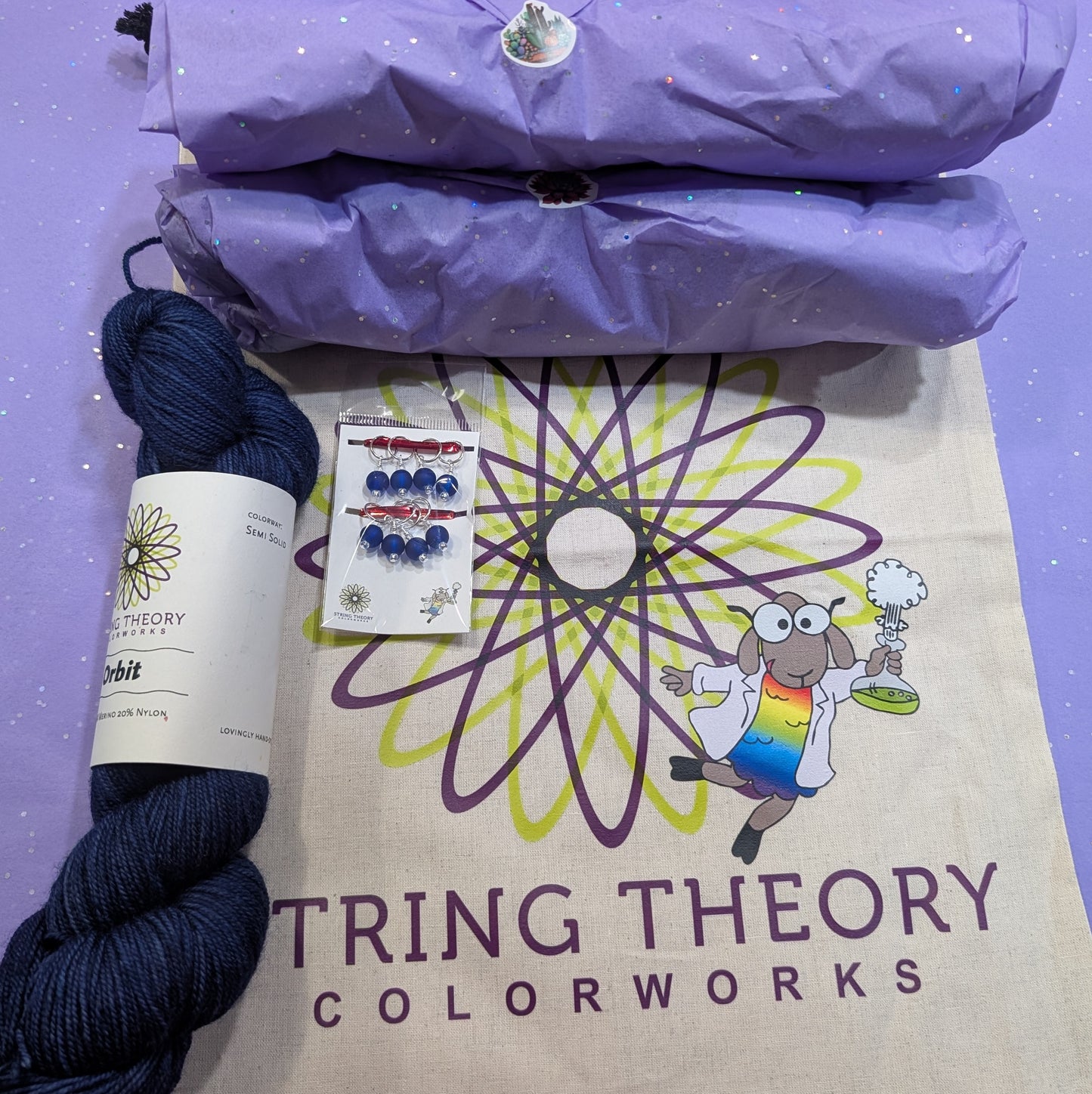 Navy yarn skein, packaging, and 'String Theory Colorworks' bag on a purple background