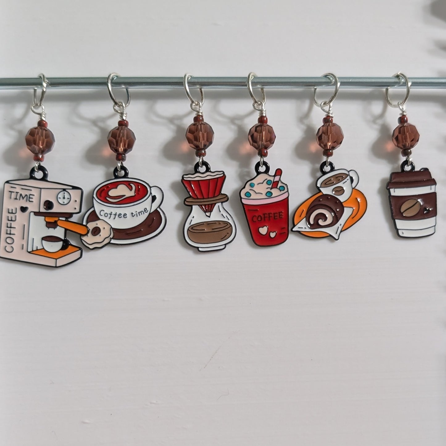 Six coffee-themed stitch markers hanging from a metal rod on a white background. The charms depict a coffee grinder, a coffee timer, a coffee cup, a pour-over dripper, a red coffee drink, and a coffee cup with a lid.