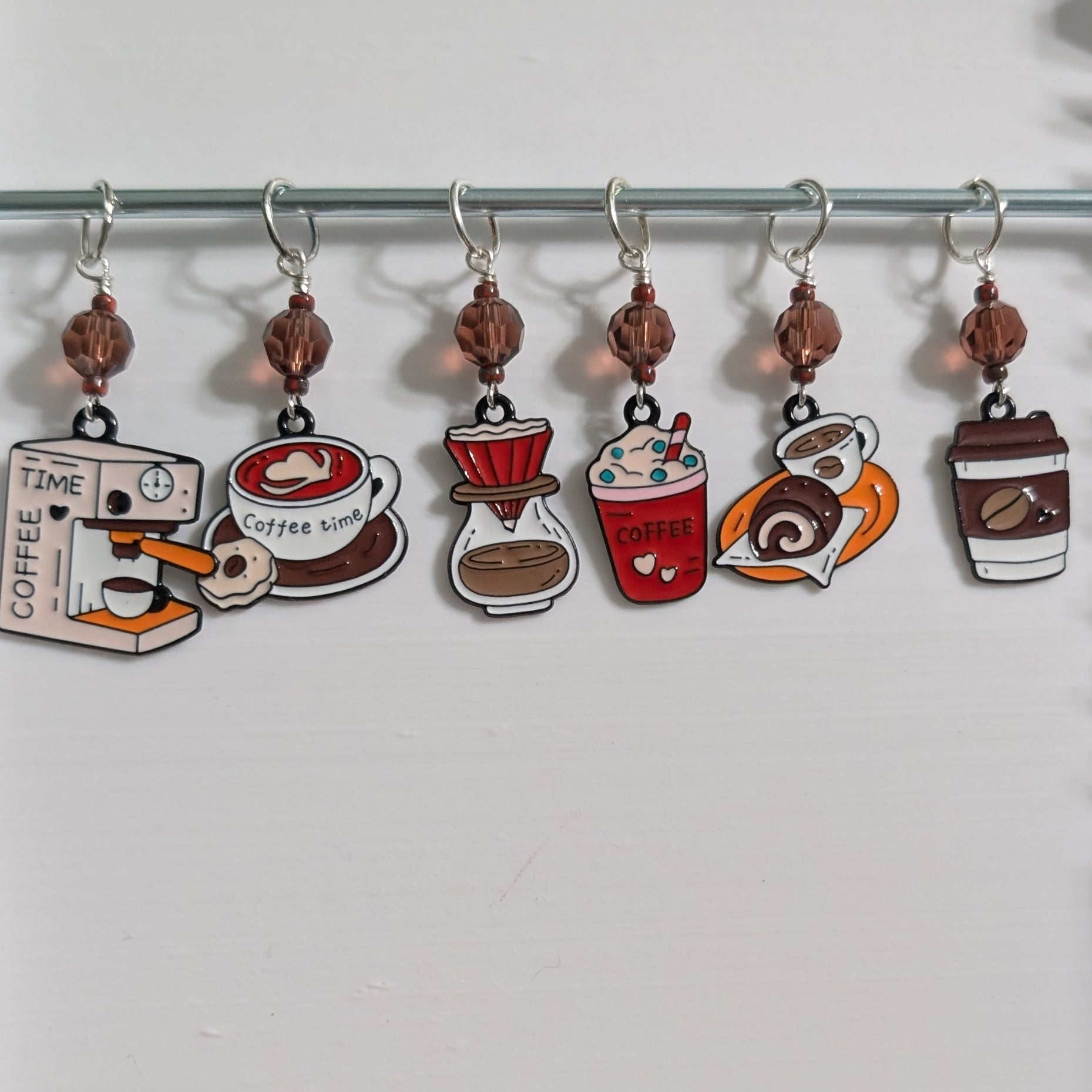 Six coffee-themed stitch markers hanging from a metal rod on a white background. The charms depict a coffee grinder, a coffee timer, a coffee cup, a pour-over dripper, a red coffee drink, and a coffee cup with a lid.