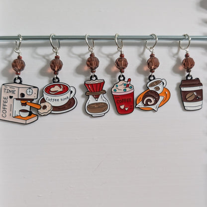 Six coffee-themed stitch markers hanging from a metal rod on a white background. The charms depict a coffee grinder, a coffee timer, a coffee cup, a pour-over dripper, a red coffee drink, and a coffee cup with a lid.