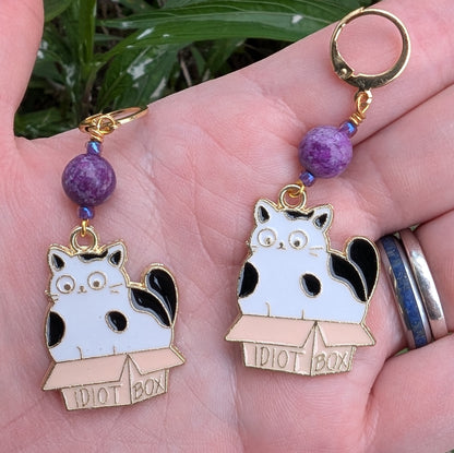 Two cat-themed earrings with purple beads held in a hand.